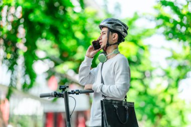 Kasklı genç bir adam, şehir caddesinde elektrikli scooterla dikilirken telefonuyla konuşuyor, modern, çevre dostu şehir ulaşımının özünü yakalıyor..