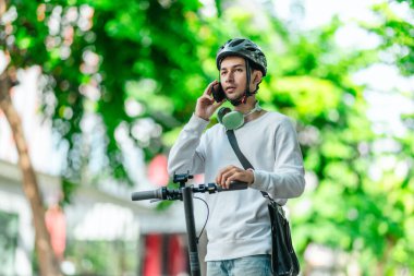 Kasklı genç bir adam, şehir caddesinde elektrikli scooterla dikilirken telefonuyla konuşuyor, modern, çevre dostu şehir ulaşımının özünü yakalıyor..