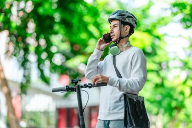 Kasklı genç bir adam, şehir caddesinde elektrikli scooterla dikilirken telefonuyla konuşuyor, modern, çevre dostu şehir ulaşımının özünü yakalıyor..