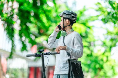 Kasklı genç bir adam, şehir caddesinde elektrikli scooterla dikilirken telefonuyla konuşuyor, modern, çevre dostu şehir ulaşımının özünü yakalıyor..