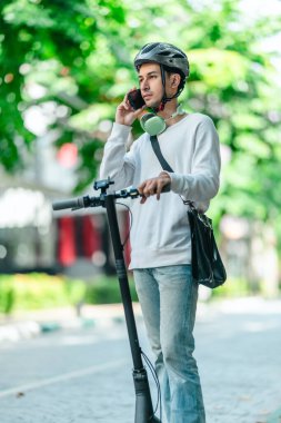 Kasklı genç bir adam, şehir caddesinde elektrikli scooterla dikilirken telefonuyla konuşuyor, modern, çevre dostu şehir ulaşımının özünü yakalıyor..