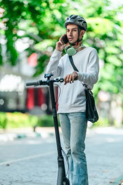 Kasklı genç bir adam, şehir caddesinde elektrikli scooterla dikilirken telefonuyla konuşuyor, modern, çevre dostu şehir ulaşımının özünü yakalıyor..