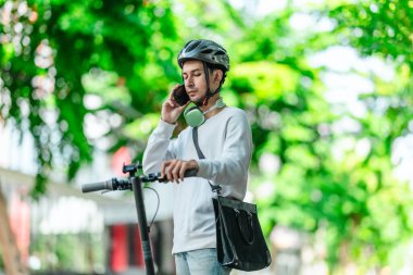 Kasklı genç bir adam, şehir caddesinde elektrikli scooterla dikilirken telefonuyla konuşuyor, modern, çevre dostu şehir ulaşımının özünü yakalıyor..