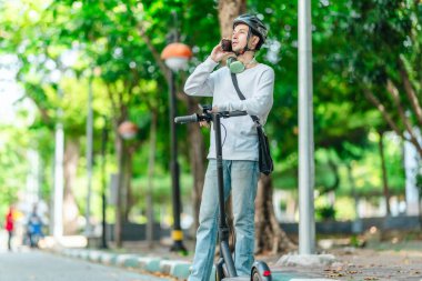 Kasklı genç bir adam, şehir caddesinde elektrikli scooterla dikilirken telefonuyla konuşuyor, modern, çevre dostu şehir ulaşımının özünü yakalıyor..