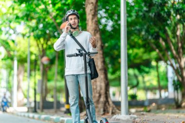 Kasklı genç bir adam, şehir caddesinde elektrikli scooterla dikilirken telefonuyla konuşuyor, modern, çevre dostu şehir ulaşımının özünü yakalıyor..