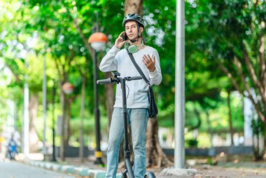 Kasklı genç bir adam, şehir caddesinde elektrikli scooterla dikilirken telefonuyla konuşuyor, modern, çevre dostu şehir ulaşımının özünü yakalıyor..