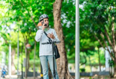 Kasklı genç bir adam, şehir caddesinde elektrikli scooterla dikilirken telefonuyla konuşuyor, modern, çevre dostu şehir ulaşımının özünü yakalıyor..