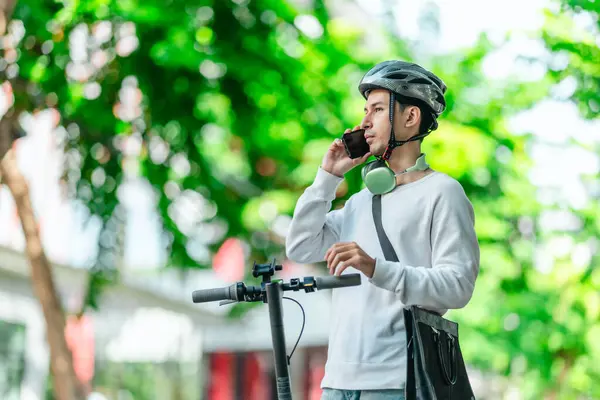Kasklı genç bir adam, şehir caddesinde elektrikli scooterla dikilirken telefonuyla konuşuyor, modern, çevre dostu şehir ulaşımının özünü yakalıyor..