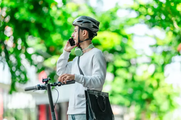 Kasklı genç bir adam, şehir caddesinde elektrikli scooterla dikilirken telefonuyla konuşuyor, modern, çevre dostu şehir ulaşımının özünü yakalıyor..