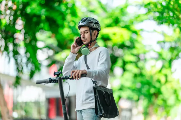 Kasklı genç bir adam, şehir caddesinde elektrikli scooterla dikilirken telefonuyla konuşuyor, modern, çevre dostu şehir ulaşımının özünü yakalıyor..