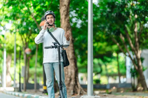 Kasklı genç bir adam, şehir caddesinde elektrikli scooterla dikilirken telefonuyla konuşuyor, modern, çevre dostu şehir ulaşımının özünü yakalıyor..
