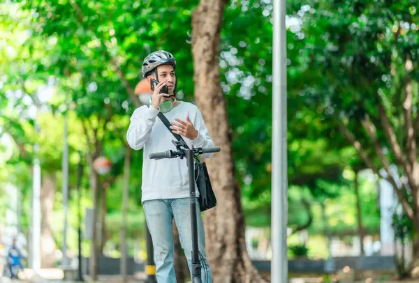 Kasklı genç bir adam, şehir caddesinde elektrikli scooterla dikilirken telefonuyla konuşuyor, modern, çevre dostu şehir ulaşımının özünü yakalıyor..