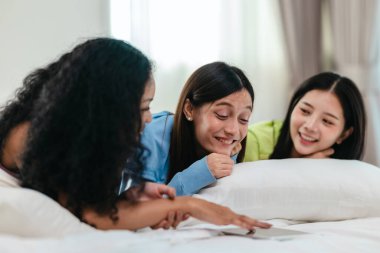 Bir grup farklı genç kadın bir yatakta uzanıyor, rahat, iyi ışıklandırılmış bir yatak odası ortamında dijital bir oyunla neşeyle meşgul oluyorlar. Görüntü arkadaşlığı, eğlenceyi ve olumlu bağlantıyı yakalıyor..