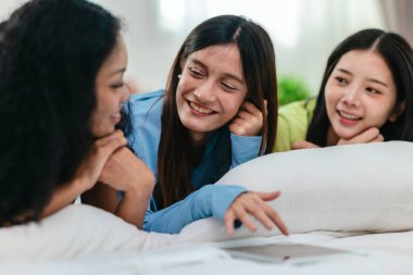 Bir grup farklı genç kadın bir yatakta uzanıyor, rahat, iyi ışıklandırılmış bir yatak odası ortamında dijital bir oyunla neşeyle meşgul oluyorlar. Görüntü arkadaşlığı, eğlenceyi ve olumlu bağlantıyı yakalıyor..