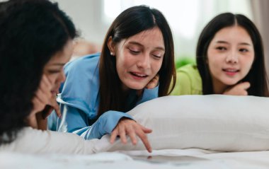 Bir grup farklı genç kadın bir yatakta uzanıyor, rahat, iyi ışıklandırılmış bir yatak odası ortamında dijital bir oyunla neşeyle meşgul oluyorlar. Görüntü arkadaşlığı, eğlenceyi ve olumlu bağlantıyı yakalıyor..
