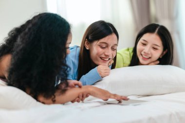 Bir grup farklı genç kadın bir yatakta uzanıyor, rahat, iyi ışıklandırılmış bir yatak odası ortamında dijital bir oyunla neşeyle meşgul oluyorlar. Görüntü arkadaşlığı, eğlenceyi ve olumlu bağlantıyı yakalıyor..