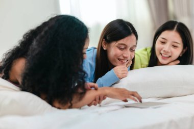 Bir grup farklı genç kadın bir yatakta uzanıyor, rahat, iyi ışıklandırılmış bir yatak odası ortamında dijital bir oyunla neşeyle meşgul oluyorlar. Görüntü arkadaşlığı, eğlenceyi ve olumlu bağlantıyı yakalıyor..