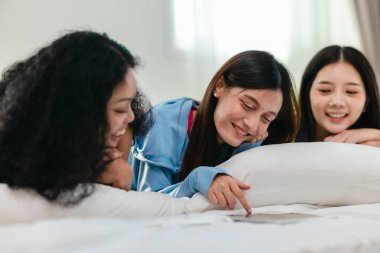 Bir grup farklı genç kadın bir yatakta uzanıyor, rahat, iyi ışıklandırılmış bir yatak odası ortamında dijital bir oyunla neşeyle meşgul oluyorlar. Görüntü arkadaşlığı, eğlenceyi ve olumlu bağlantıyı yakalıyor..