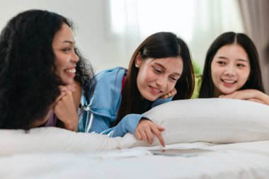 Bir grup farklı genç kadın bir yatakta uzanıyor, rahat, iyi ışıklandırılmış bir yatak odası ortamında dijital bir oyunla neşeyle meşgul oluyorlar. Görüntü arkadaşlığı, eğlenceyi ve olumlu bağlantıyı yakalıyor..