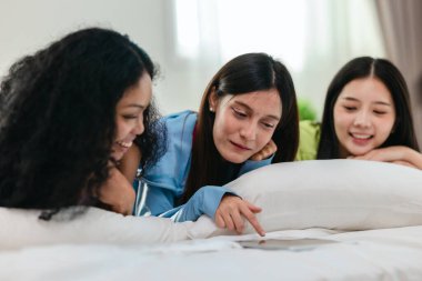 Bir grup farklı genç kadın bir yatakta uzanıyor, rahat, iyi ışıklandırılmış bir yatak odası ortamında dijital bir oyunla neşeyle meşgul oluyorlar. Görüntü arkadaşlığı, eğlenceyi ve olumlu bağlantıyı yakalıyor..