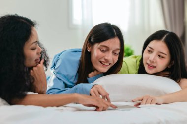 Bir grup farklı genç kadın bir yatakta uzanıyor, rahat, iyi ışıklandırılmış bir yatak odası ortamında dijital bir oyunla neşeyle meşgul oluyorlar. Görüntü arkadaşlığı, eğlenceyi ve olumlu bağlantıyı yakalıyor..