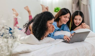 Bir grup farklı genç kadın bir yatakta uzanıyor, rahat, iyi ışıklandırılmış bir yatak odası ortamında dijital bir oyunla neşeyle meşgul oluyorlar. Görüntü arkadaşlığı, eğlenceyi ve olumlu bağlantıyı yakalıyor..