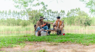 İki Asyalı erkek arkadaş, sakin bir göl kenarında, piknik sırasında akıllı telefonu şarj etmek için taşınabilir bir güneş paneli kullanıyorlar. Sahne, doğa ve modern sürdürülebilir yaşamın bir karışımını yansıtıyor..