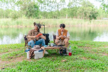 İki Asyalı erkek arkadaş, sakin bir göl kenarında, piknik sırasında akıllı telefonu şarj etmek için taşınabilir bir güneş paneli kullanıyorlar. Sahne, doğa ve modern sürdürülebilir yaşamın bir karışımını yansıtıyor..