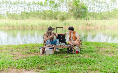 İki Asyalı erkek arkadaş, sakin bir göl kenarında, piknik sırasında akıllı telefonu şarj etmek için taşınabilir bir güneş paneli kullanıyorlar. Sahne, doğa ve modern sürdürülebilir yaşamın bir karışımını yansıtıyor..