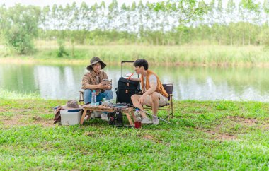 İki Asyalı erkek arkadaş, sakin bir göl kenarında, piknik sırasında akıllı telefonu şarj etmek için taşınabilir bir güneş paneli kullanıyorlar. Sahne, doğa ve modern sürdürülebilir yaşamın bir karışımını yansıtıyor..