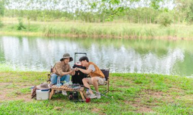 İki Asyalı erkek arkadaş, sakin bir göl kenarında, piknik sırasında akıllı telefonu şarj etmek için taşınabilir bir güneş paneli kullanıyorlar. Sahne, doğa ve modern sürdürülebilir yaşamın bir karışımını yansıtıyor..