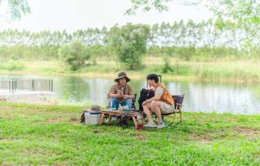 İki Asyalı erkek arkadaş, sakin bir göl kenarında, piknik sırasında akıllı telefonu şarj etmek için taşınabilir bir güneş paneli kullanıyorlar. Sahne, doğa ve modern sürdürülebilir yaşamın bir karışımını yansıtıyor..