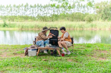 İki Asyalı erkek arkadaş, sakin bir göl kenarında, piknik sırasında akıllı telefonu şarj etmek için taşınabilir bir güneş paneli kullanıyorlar. Sahne, doğa ve modern sürdürülebilir yaşamın bir karışımını yansıtıyor..
