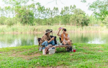 İki Asyalı erkek arkadaş, sakin bir göl kenarında, piknik sırasında akıllı telefonu şarj etmek için taşınabilir bir güneş paneli kullanıyorlar. Sahne, doğa ve modern sürdürülebilir yaşamın bir karışımını yansıtıyor..