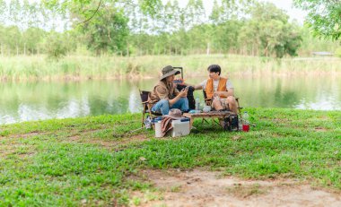 İki Asyalı erkek arkadaş, sakin bir göl kenarında, piknik sırasında akıllı telefonu şarj etmek için taşınabilir bir güneş paneli kullanıyorlar. Sahne, doğa ve modern sürdürülebilir yaşamın bir karışımını yansıtıyor..