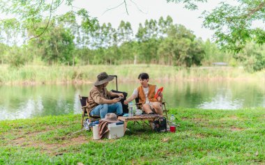 İki Asyalı erkek arkadaş, sakin bir göl kenarında, piknik sırasında akıllı telefonu şarj etmek için taşınabilir bir güneş paneli kullanıyorlar. Sahne, doğa ve modern sürdürülebilir yaşamın bir karışımını yansıtıyor..