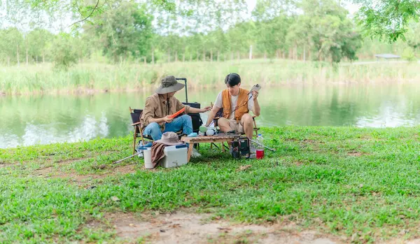 İki Asyalı erkek arkadaş, sakin bir göl kenarında, piknik sırasında akıllı telefonu şarj etmek için taşınabilir bir güneş paneli kullanıyorlar. Sahne, doğa ve modern sürdürülebilir yaşamın bir karışımını yansıtıyor..