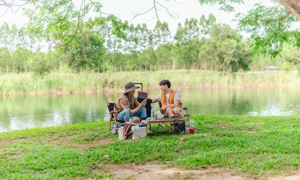 İki Asyalı erkek arkadaş, sakin bir göl kenarında, piknik sırasında akıllı telefonu şarj etmek için taşınabilir bir güneş paneli kullanıyorlar. Sahne, doğa ve modern sürdürülebilir yaşamın bir karışımını yansıtıyor..