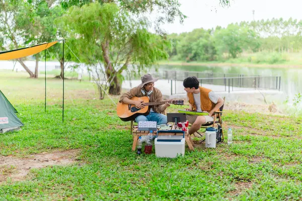 İki Asyalı erkek arkadaş gölün kenarında dinleniyor, müziğin tadını çıkarıyor ve açık hava ortamında birlikte şarkı söylüyorlar. Sahne, kamp gezisi sırasında neşeli ve huzurlu bir bağ ve yaratıcılık anı yakalıyor.