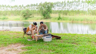 İki genç Asyalı erkek, kamp alanında dinlenirken tablet üzerinde video oyunu oynamaktan hoşlanırlar. Kamp malzemeleri, gitarlar ve doğa ile çevrili sahne, modern ve neşeli doğa deneyimini yakalar..