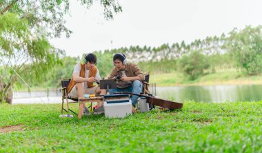 İki genç Asyalı erkek, kamp alanında dinlenirken tablet üzerinde video oyunu oynamaktan hoşlanırlar. Kamp malzemeleri, gitarlar ve doğa ile çevrili sahne, modern ve neşeli doğa deneyimini yakalar..