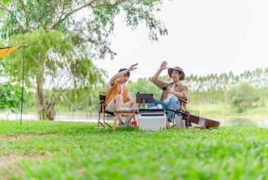 İki genç Asyalı erkek, kamp alanında dinlenirken tablet üzerinde video oyunu oynamaktan hoşlanırlar. Kamp malzemeleri, gitarlar ve doğa ile çevrili sahne, modern ve neşeli doğa deneyimini yakalar..