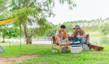 İki genç Asyalı erkek, kamp alanında dinlenirken tablet üzerinde video oyunu oynamaktan hoşlanırlar. Kamp malzemeleri, gitarlar ve doğa ile çevrili sahne, modern ve neşeli doğa deneyimini yakalar..
