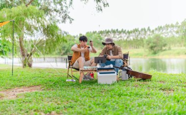 İki genç Asyalı erkek, kamp alanında dinlenirken tablet üzerinde video oyunu oynamaktan hoşlanırlar. Kamp malzemeleri, gitarlar ve doğa ile çevrili sahne, modern ve neşeli doğa deneyimini yakalar..