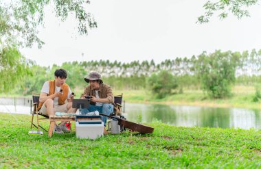 İki genç Asyalı erkek, kamp alanında dinlenirken tablet üzerinde video oyunu oynamaktan hoşlanırlar. Kamp malzemeleri, gitarlar ve doğa ile çevrili sahne, modern ve neşeli doğa deneyimini yakalar..