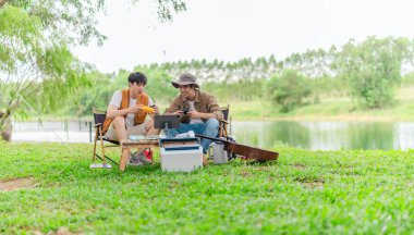 İki genç Asyalı erkek, kamp alanında dinlenirken tablet üzerinde video oyunu oynamaktan hoşlanırlar. Kamp malzemeleri, gitarlar ve doğa ile çevrili sahne, modern ve neşeli doğa deneyimini yakalar..