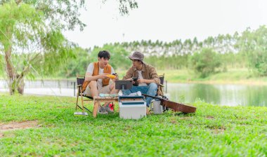 İki genç Asyalı erkek, kamp alanında dinlenirken tablet üzerinde video oyunu oynamaktan hoşlanırlar. Kamp malzemeleri, gitarlar ve doğa ile çevrili sahne, modern ve neşeli doğa deneyimini yakalar..