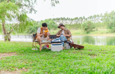 İki genç Asyalı erkek, kamp alanında dinlenirken tablet üzerinde video oyunu oynamaktan hoşlanırlar. Kamp malzemeleri, gitarlar ve doğa ile çevrili sahne, modern ve neşeli doğa deneyimini yakalar..
