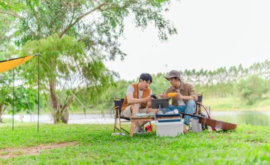 İki genç Asyalı erkek, kamp alanında dinlenirken tablet üzerinde video oyunu oynamaktan hoşlanırlar. Kamp malzemeleri, gitarlar ve doğa ile çevrili sahne, modern ve neşeli doğa deneyimini yakalar..
