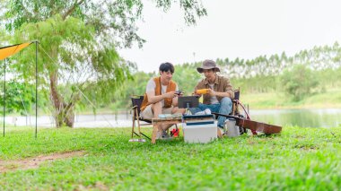 İki genç Asyalı erkek, kamp alanında dinlenirken tablet üzerinde video oyunu oynamaktan hoşlanırlar. Kamp malzemeleri, gitarlar ve doğa ile çevrili sahne, modern ve neşeli doğa deneyimini yakalar..